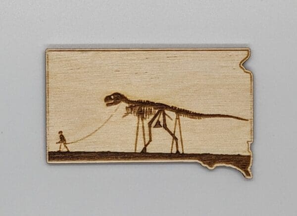 Custom wood magnet in the shape of South Dakota depicting a skeleton man walking a skeleton dinosaur